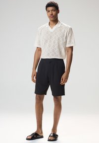 Krém színű rövid ujjú kötött galléros póló gyémántmintával; fekete redős rövidnadrág; fekete papucs szandál. Egyszerű, kortárs design.