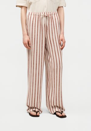 Oseba nosi široke hlače bež in rdeče navpično črtaste, z vrvico v pasu in črnimi natikači, stoji pred belim ozadjem.