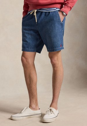 Muž oblečený v modrých riflových šortkách so sťahovacou šnúrkou, biele tenisky a červená mikina, stojaci s rukami v vreckách.