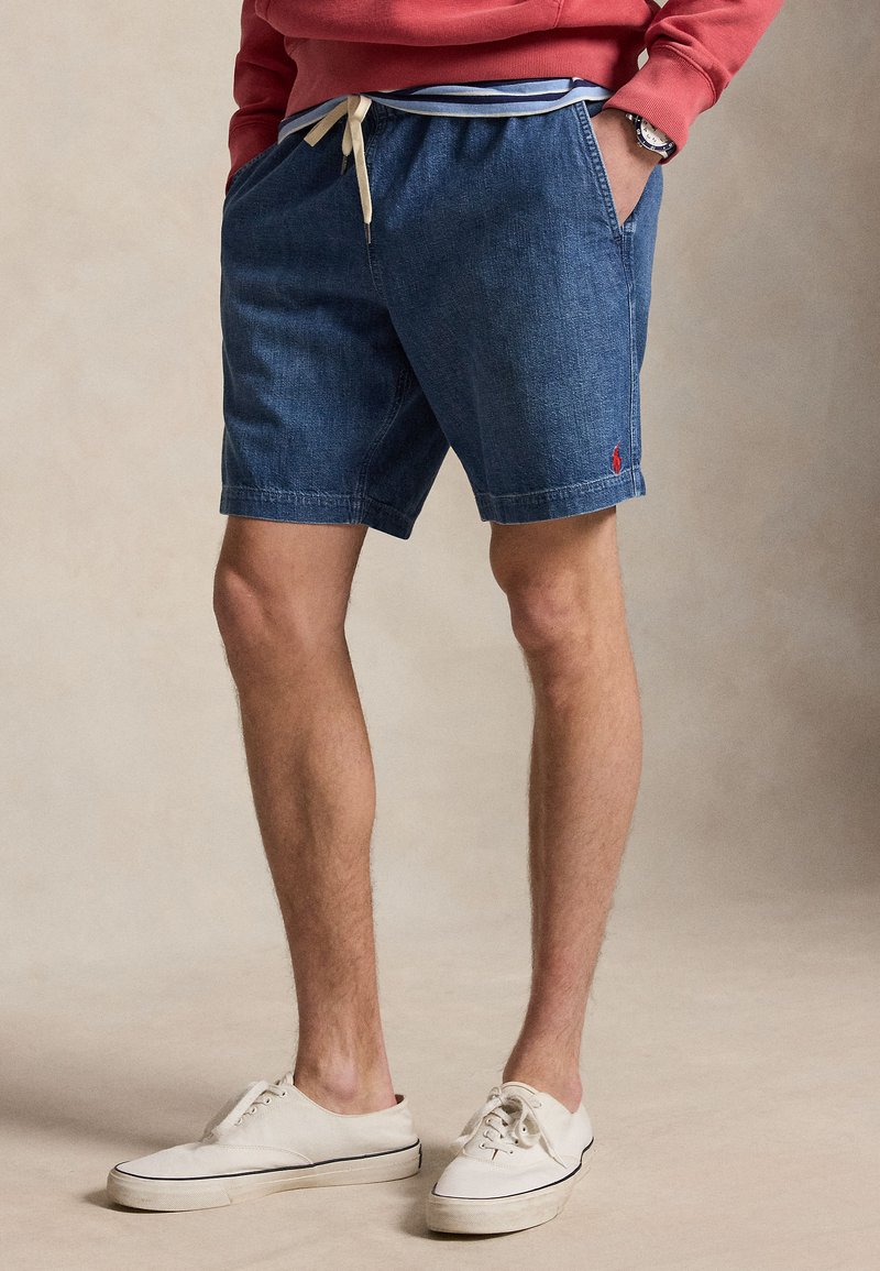 Άνδρας με μπλε τζιν σορτς με κορδόνι, λευκά αθλητικά παπούτσια και κόκκινο φούτερ, στέκεται με τα χέρια στις τσέπες.