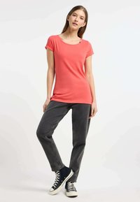 T-shirt corallo a maniche corte con scollo rotondo e dettaglio decorativo a bottoni, abbinato a pantaloni in denim grigi e sneaker scure.