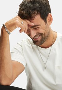 Zilveren ketting met een rechthoekige hanger, gedragen over een simpele witte T-shirt. Een zilveren ring en armband zijn ook zichtbaar.