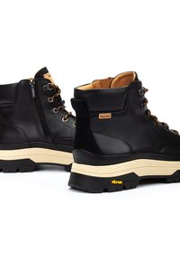Botas de senderismo de cuero negro con suelas de Vibram beige, con cordones en la parte delantera, cremallera lateral y forro interior beige; cuenta con ojales metálicos y acentos con el logotipo.