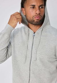 Sweat à capuche gris clair avec fermeture éclair sur le devant, capuche réglable avec cordon de serrage, et poignets côtelés. Confectionné en tissu doux avec une texture lisse.