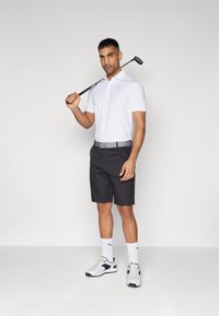 Bílé polokošile, černé šortky, šedý opasek a černá golfová hůl. Bílé sportovní boty s černými akcenty. Postoj ve stoje na jednobarevném pozadí.