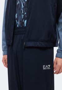 Άτομο που φοράει σκούρες μπλε παντελόνια EA7 Emporio Armani, ένα πουκάμισο με σχέδια και ένα σκούρο μπλε και ανοιχτό μπλε μπουφάν με ελαστικές μανσέτες.
