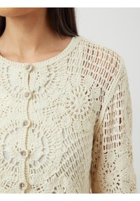 Szydełkowy kardigan w kolorze off-white, z kwiatowymi wzorami, okrągłym dekoltem i perłowymi guzikami. Otwarta struktura splotu, z długimi rękawami.