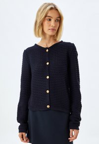 Marinblå stickad cardigan med en texturerad mönster, rund halsringning och guldfärgad knappar. Långa ärmar med ribbade manschetter. Avslappnad passform.