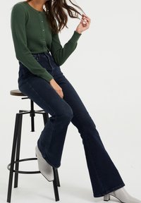 Grön texturerad cardigan med knappar, kombinerad med högmidjade mörkblå bootcut-jeans och ljusgrå ankelstövlar, sittande på en pall.