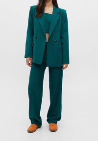 Turkusowy oversize'owy żakiet z podwójnymi guzikami, dopasowany turkusowy krótki top i luźne spodnie. Noszony z pomarańczowymi zamszowymi sneakersami.