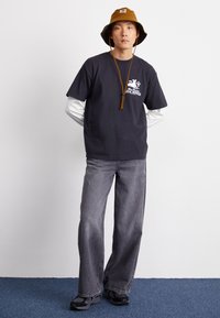 Dunkelgraues T-Shirt mit Grafikdruck, kombiniert mit weiten grauen Jeans. Ein brauner Bucket Hat sitzt obenauf, und schwarze Sneaker runden den Look ab.