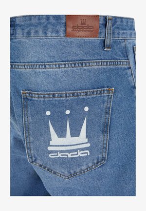 Sinine denimist teksapüksid, millel on tagaküljel tasku, millel on valge krooni graafika ja logo "dada" allpool. Nahkline vööpõhi on kaasas.