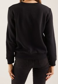 Felpa nera in cotone con collo rotondo, polsini e fondo a costine. Abbinata a jeans scuri, che mostrano una vestibilità slim e dettagli minimi.