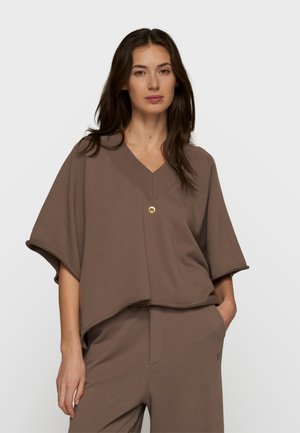 Donna con lunghi capelli scuri che indossa una maglia marrone scuro a V con maniche corte e pantaloni coordinati, in piedi con una mano in tasca.