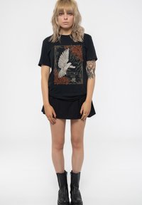 Black t-shirt featuring a graphic of a white bird, surrounded by red foliage and gold text, paired with a black pleated skirt and black boots.