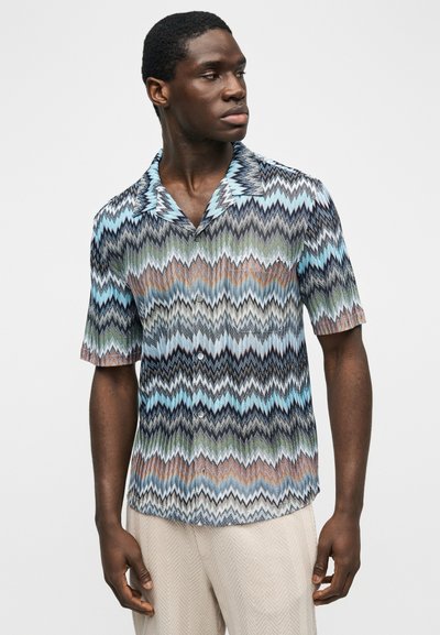 Άνδρας που φοράει κοντομάνικο πουκάμισο με σχέδιο ζιγκ-ζαγκ σε μπλε, μαύρο, πράσινο και καφέ, σε συνδυασμό με ανοιχτόχρωμο παντελόνι με υφή.