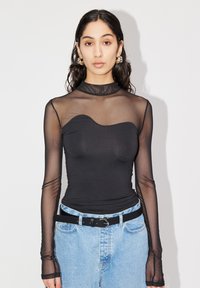 Body in rete nero con maniche lunghe, colletto alto e scollatura asimmetrica. Abbinato a jeans azzurri a vita alta e una cintura nera.