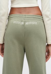 Γυναίκα που φοράει χακί φόρμα Nike Shox και κρεμ μακρυμάνικη μπλούζα, εμφανιζόμενη από τη μέση έως το μέσο του μηρού, σε πλάγια όψη από πίσω.