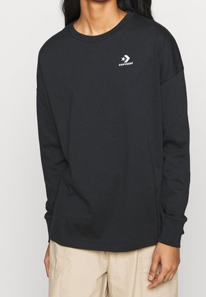 Persoon draagt een zwart langarmig Converse-shirt met logo op de borst, gecombineerd met lichtbeige broek, staand tegen een effen achtergrond.