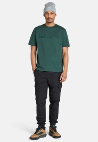 Hombre con camiseta verde de Timberland, gorro gris, pantalones de cargo negros y botas de senderismo marrones, de pie con las manos en los bolsillos sobre un fondo blanco.