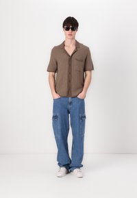Cămașă scurtă, cu mânecă, de culoare maro, cu un model texturat și buzunar pe piept, asortată cu blugi cargo bleumarin deschis și adidași albi.