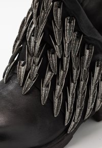 Svart läderstövel med metalliska taggar som är arrangerade i ett lager-på-lager-mönster, med texturerade detaljer och en rund tådesign.