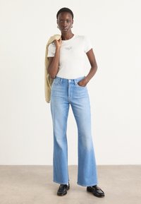Jeans flared di un blu chiaro in denim, a vita alta, abbinati a una t-shirt bianca attillata con testo piccolo. Il modello tiene in mano un giubbotto beige.