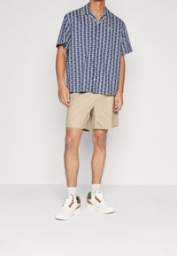 Košile s krátkým rukávem, zapínání na knoflíky v námořnické modré barvě s geometrickým bílým vzorem, zkombinována s světle béžovými šortkami a bílými teniskami s tanovými akcenty.