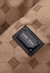 Hnědá textilní tkanina s embossovaným vzorem loga. Obsahuje černou etiketu se zněním "OMBRA EXIMIO" bílým písmem, upevněnou čtyřmi kovovými nýty.