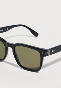 Črna pravokotna sončna očala z zelenkasto obarvanimi lečami in majhnim belim logotipom krokodila na ročaju, na svetli podlagi.