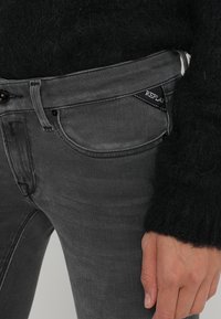 Persona que lleva pantalones oscuros de mezclilla con una etiqueta de REPLAY visible en el bolsillo, un suéter negro de felpa y la mano apoyada cerca del muslo.