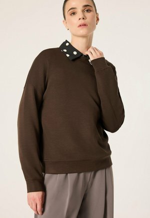 Femme portant un pull marron foncé par-dessus une chemise noire à pois blancs, associée à un pantalon gris ample, sur un fond clair uni.