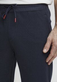 Jūras spēku zilas sporta bikses ar elastīgu jostasvietu, auklu ar sarkaniem akcentiem, un gludu tekstūru. Tuvplāns uz kabatas zonu.