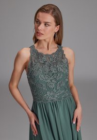 Fließendes Abendkleid in gedämpftem Grün, mit einem floralen Spitzenoberteil mit kunstvoller Perlenbestickung und gewelltem Ausschnitt, weichem Stoff und plissiertem Rock.