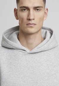 Gaišs pelēks hoody ar mīkstu, teksturētu audumu, kuram ir liela, izteikta kapuce un vienkāršs dizains. Tuvplāns skats uz kakla apkakles zonu.