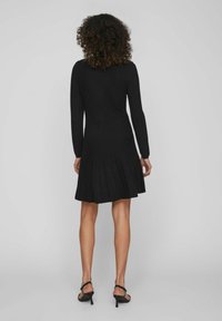 Donna con capelli ricci che indossa un vestito lungo a maniche lunghe a coste nero e sandali neri con tacco, in piedi contro uno sfondo grigio neutro.