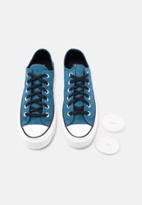 Ζευγάρι μπλε χαμηλά αθλητικά παπούτσια με μαύρα κορδόνια και λευκές λαστιχένιες μύτες, συνοδευόμενα από δύο επιπλέον ζευγάρια λευκών κορδονιών σε λευκό φόντο.