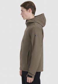 Jeune homme de profil portant une veste à capuche taupe avec logo 4F sur la manche, debout devant un fond clair uni.