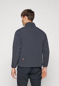 Marineblå pufferjakke med høy krage, strukturert stoff og en baklomme med oransjefarget knappdetalj. Enkel og funksjonell design.