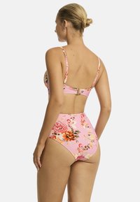 Modelka w różowym bikini w kwiaty, odwrócona, eksponująca wysokie majtki i odkryty tył, na tle jednolitego białego tła.