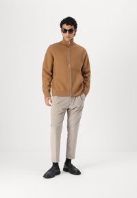 Brun zip-up jakke i et mykt materiale, grå stripete bukser, svarte sko og solbriller, med en avslappet passform og minimalistisk design.