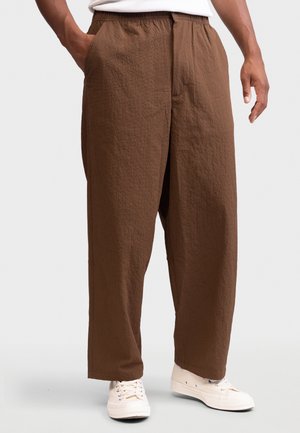 Homme portant un pantalon marron ample texturé et des baskets blanches, debout avec une main dans la poche et l'autre détendue le long du corps.