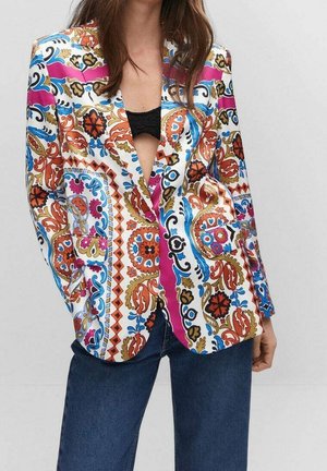 Kvinde iført en farverig blazer med blomstermønster og geometrisk design over en sort top og blå jeans mod en ensfarvet baggrund.
