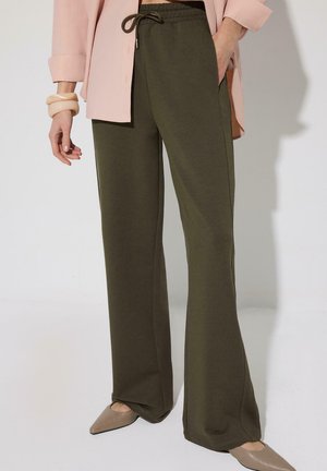 Kvinde iført olivengrønne vidde bukser med snøre, rosa blazer, beige spidse flade sko og chunky beige armbånd.