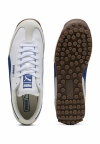 Baskets blanches et marine avec un dessus en daim et cuir, présentant un design à lacets, une semelle en caoutchouc avec des clous de grip circulaires et des accents de logo.