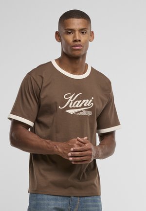 Jeune homme aux cheveux courts portant une chemise marron à manches courtes avec bordure blanche et le texte "Kani", debout, les mains jointes.