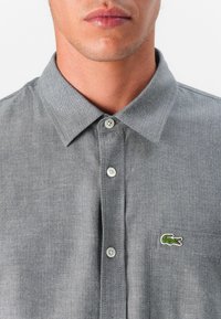Camisa de botones en espiga gris con cuello puntiagudo, que presenta botones blancos y un pequeño logo de cocodrilo verde en el lado izquierdo del pecho.