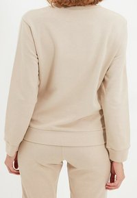 Persona che indossa un top beige a maniche lunghe e pantaloni abbinati, mostrata di spalle su uno sfondo neutro.
