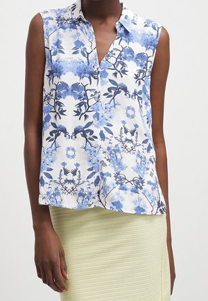 Blusa blanca sin mangas con estampado azul de flores y pájaros, combinada con una falda de canalé de color amarillo claro, llevada por una persona que está de pie frente a un fondo liso.