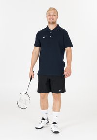 Marinblå pikétröja med FZ-logga, svarta shorts, vita sportstrumpor och svartvita sneakers, hållande en badmintonracket.